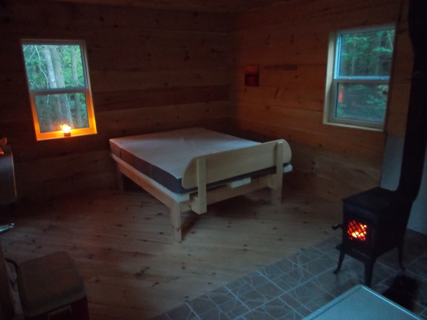 Inside cabin at dusk, a queen-size bed is lit by a warm woodstove, and an orange electric lantern